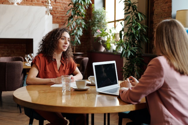 Woman sitting on table with cups of coffee and laptop while talking to another person